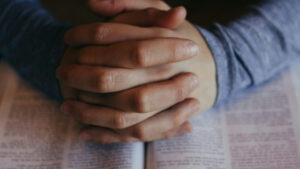 A man's praying hands rest on an open Bible; Patrick Fore / Unsplash