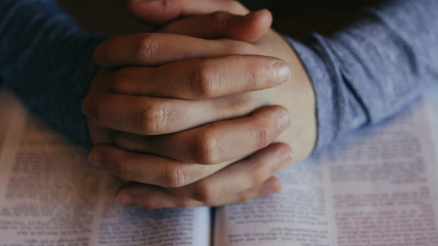 A man's praying hands rest on an open Bible; Patrick Fore / Unsplash