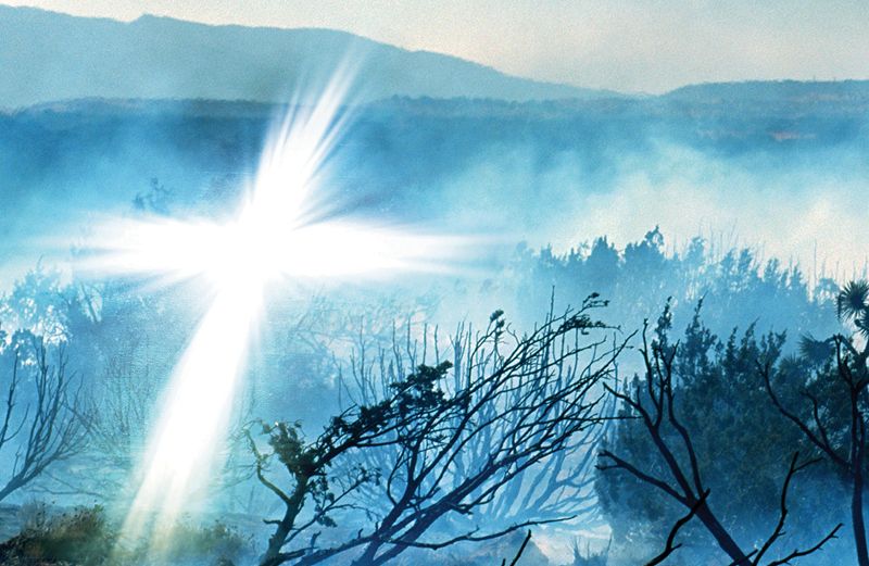 A glowing white cross appears in a smoky, fire-ravaged landscape.