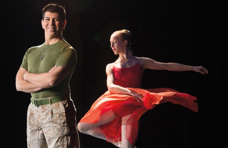 Roman stands in a spotlight, while a ballerina dances a few feet behind.