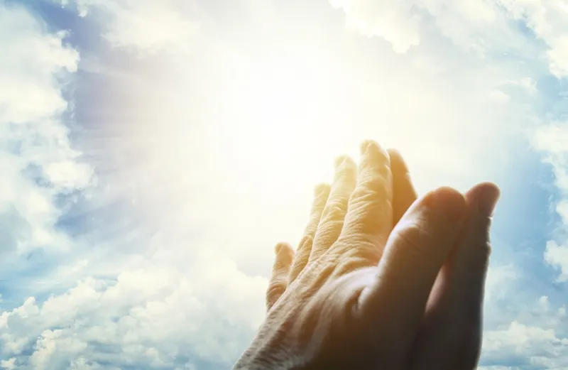 Praying hands lifted to a sunlit sky.