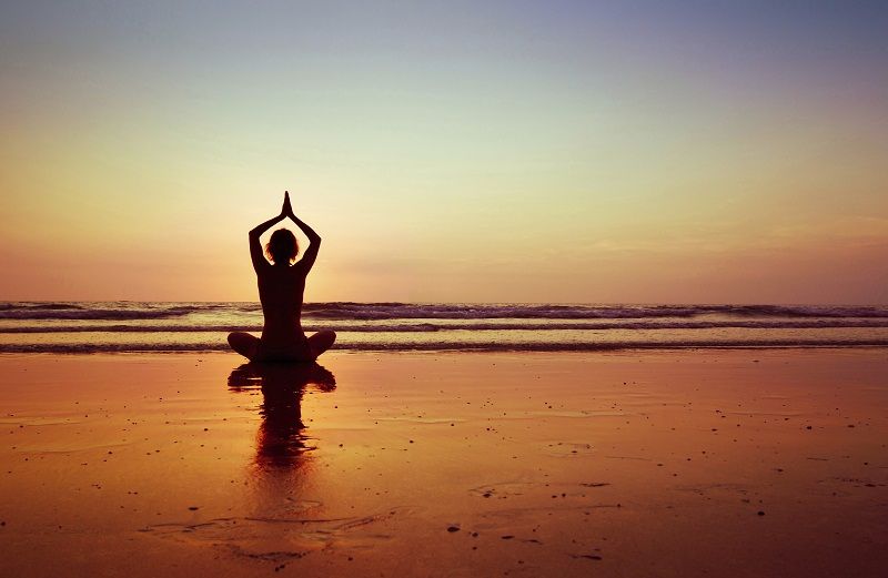 A woman sits on a beach doing yoga in front of the sunset