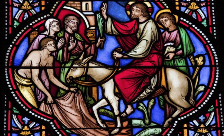 Stained Glass image of Jesus riding a donkey before a crowd laying palms on Palm Sunday