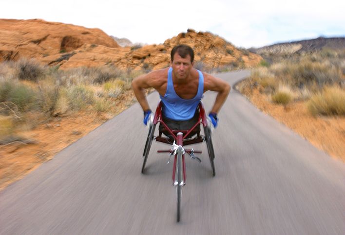 A man in a racing wheelchair sprints along a desert road.