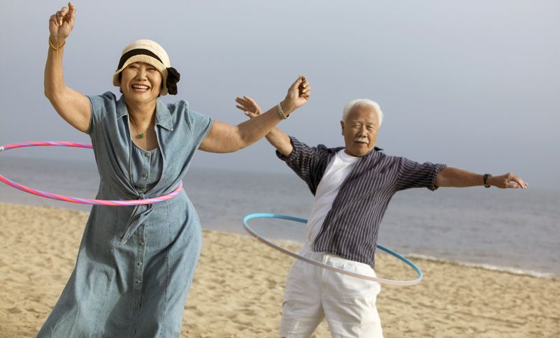 A woman and her father laugh as they hula hoop on the beach.