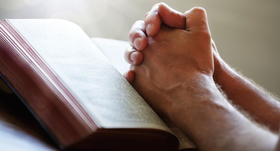 A man's praying hands rest upon an open Bible.
