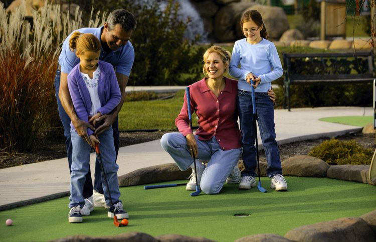 A father shows his daughter how to putt on a mini-golf course as Mom and brother look on.