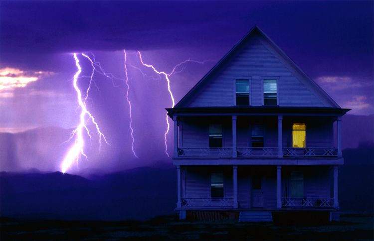 A huge flash of lightning appears behind an inviting rural home, lit from within.
