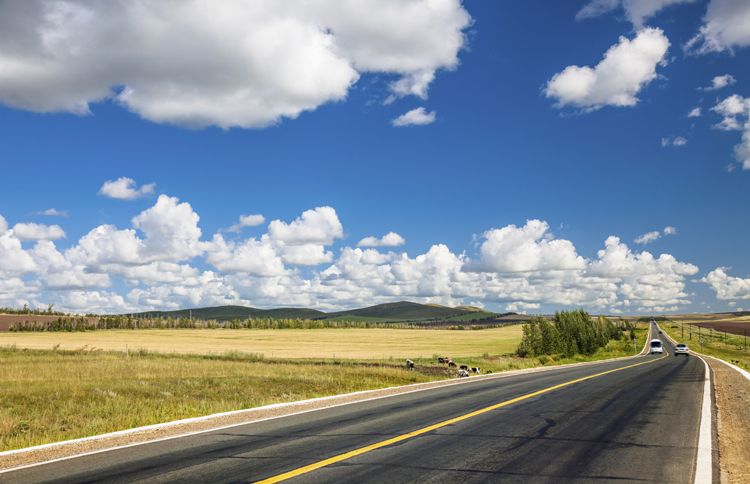 Puffy clouds in a bright blue sky hover over a beckoning highway that stretches into the distance.