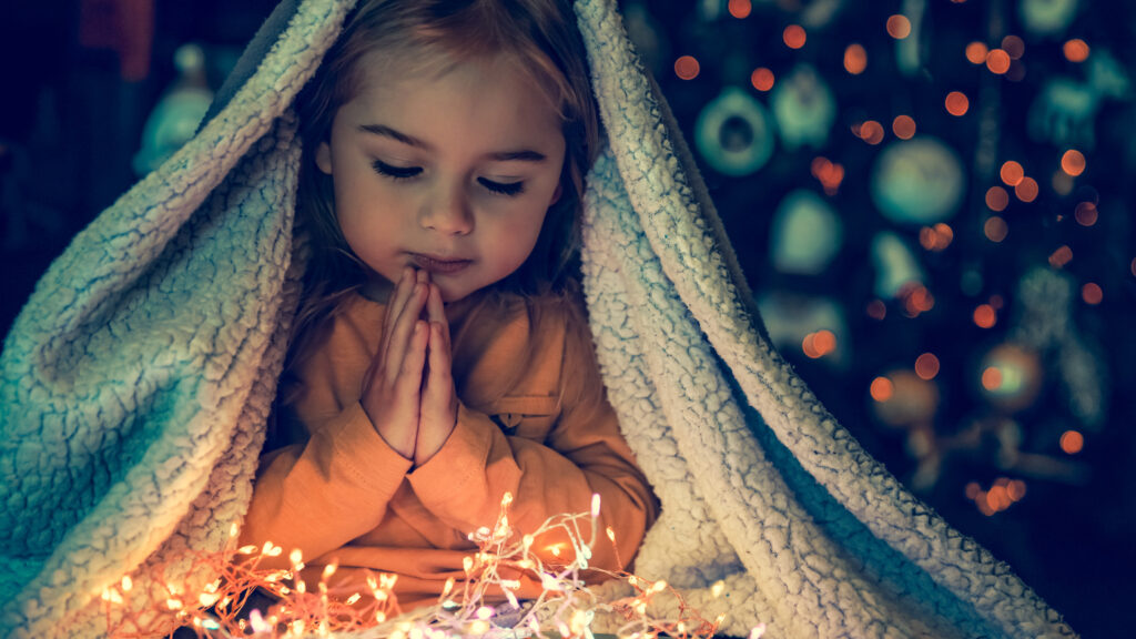 A little girl under a blanket says a christmas prayer while looking at lights