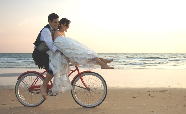 A bride and groom laugh about their wedding blessings while riding a bike on the beach