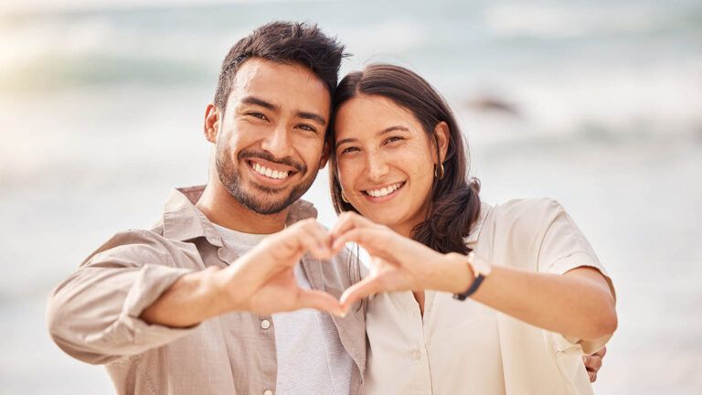 Young newlywed couple on the beach making a heart with their hands