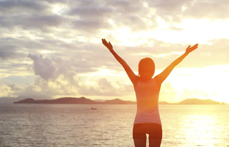 A woman stands on the beach at dawn with her arms raised in praise