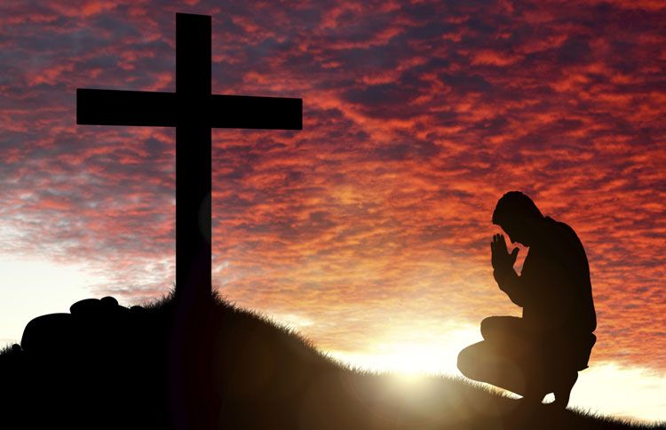 A man kneels at the foot of a cross on a hill at sunset