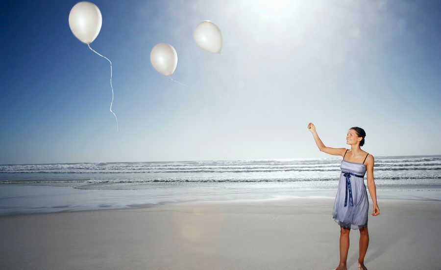 A smiling woman sets free a trio of white balloons on a sun-drenched beach