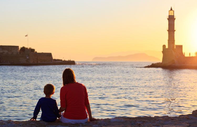 A mother and son sit on a beach at sunset and gaze at a lighthouse