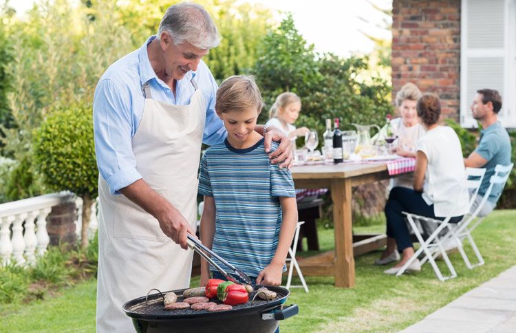 A grandfather teaches his grandson about grilling outdoors while the family looks on