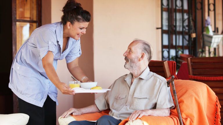 a nurse with a tray of food bends over to offer it to an elderly man sitting in a chair