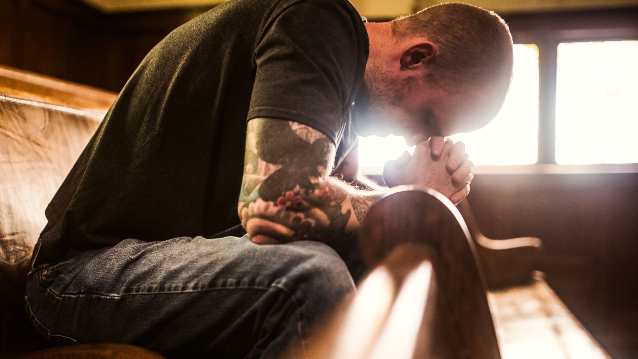 a man in a short-sleeved shirt with tattoos is sitting on a pew, bent over in prayer