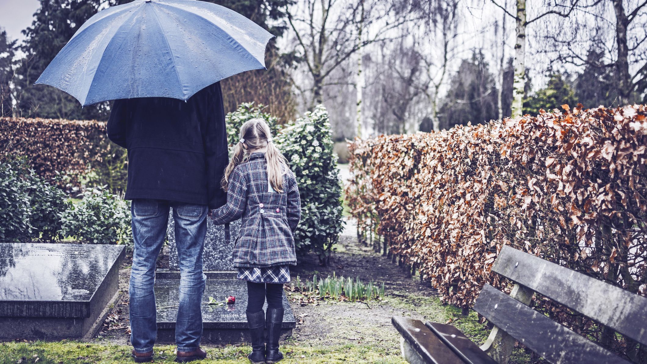 a father and a young daughter hold hands in the rain under an umbrella, standing over a grave