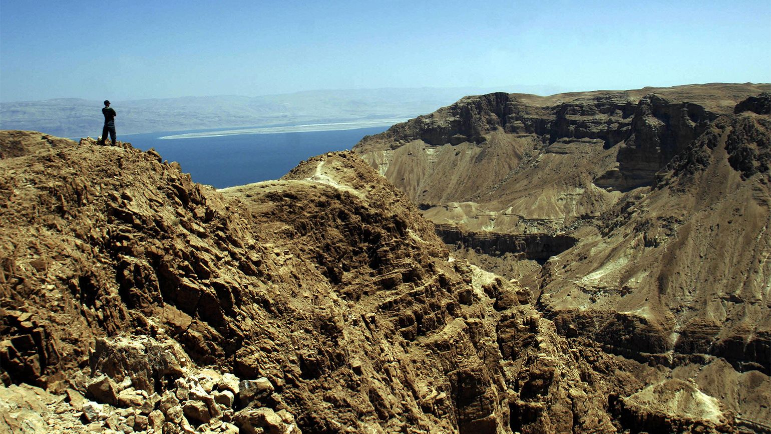 A lone figure stands upon the jagged rocks of Qumran and gazes out over the Dead Sea