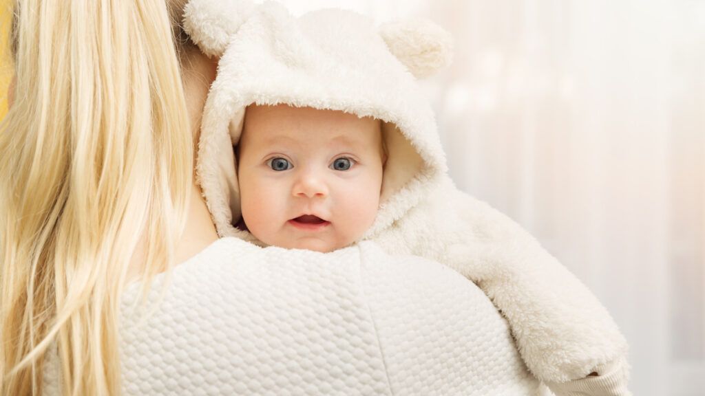 A happy baby in a warm, fuzzy polar bear jumpsuit held up by his mother's arms.