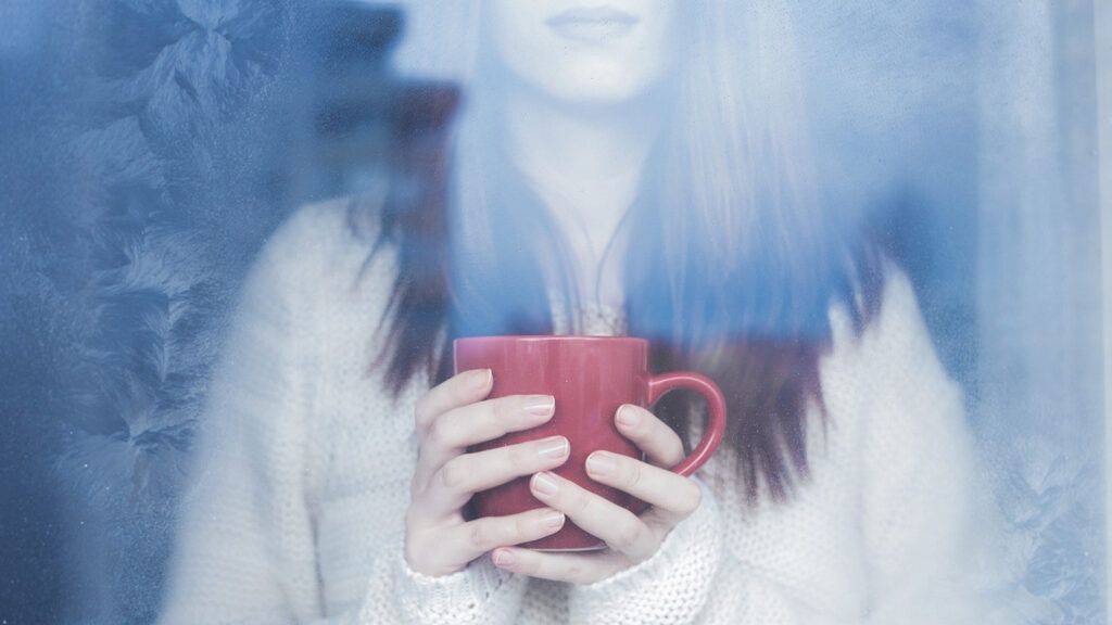 A woman gazes out a window, a warm cup of tea cradled in her hands