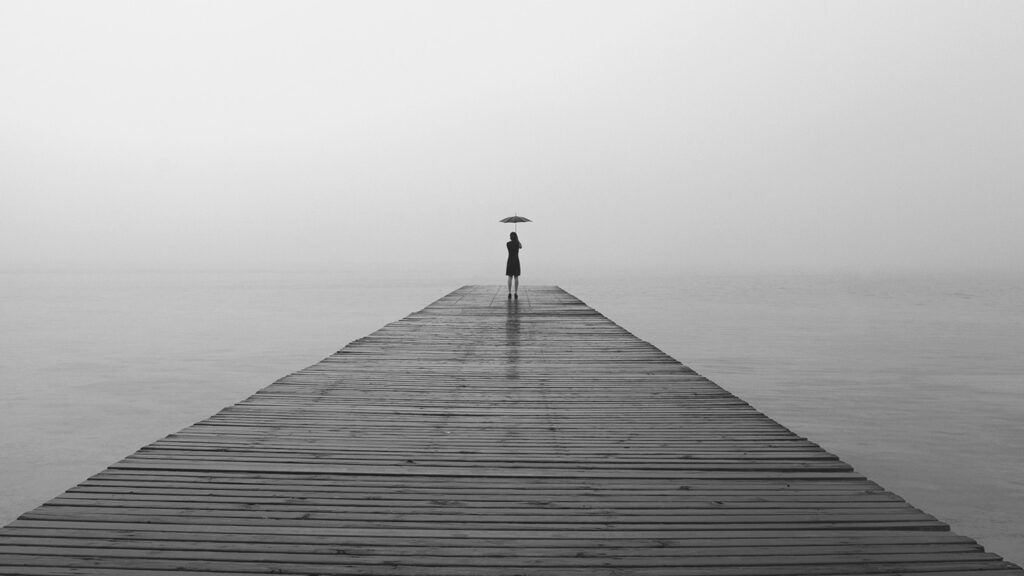 A woman stands at the end of a long, foggy pier, umbrella in hand