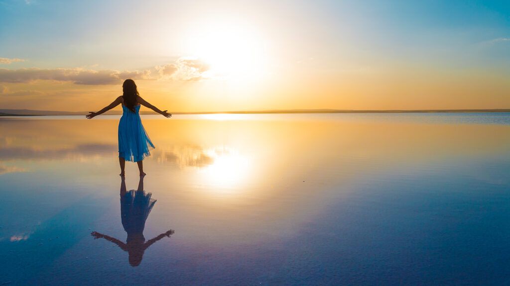 A woman stands at the edge of still water that reflects a sunrise