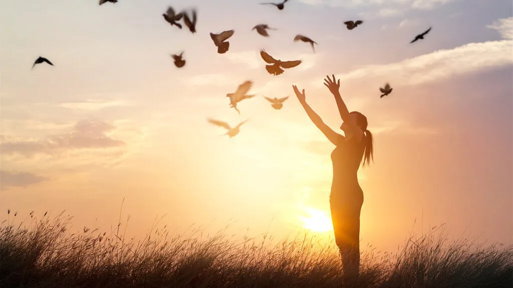 A woman with arms upraised at sunrise, a flock of doves flying around her