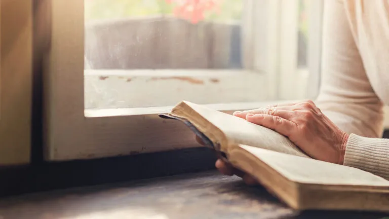 A woman reads her Bible while seated by a window where the sun shines in