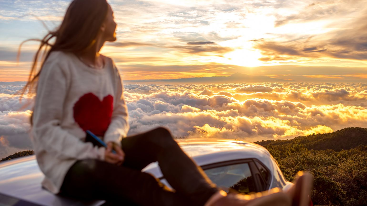 Young woman in sweater with heart shape enjoying beautiful cloudscape sitting on the car roof above the clouds on the sunrise. Image focused on the clouds
