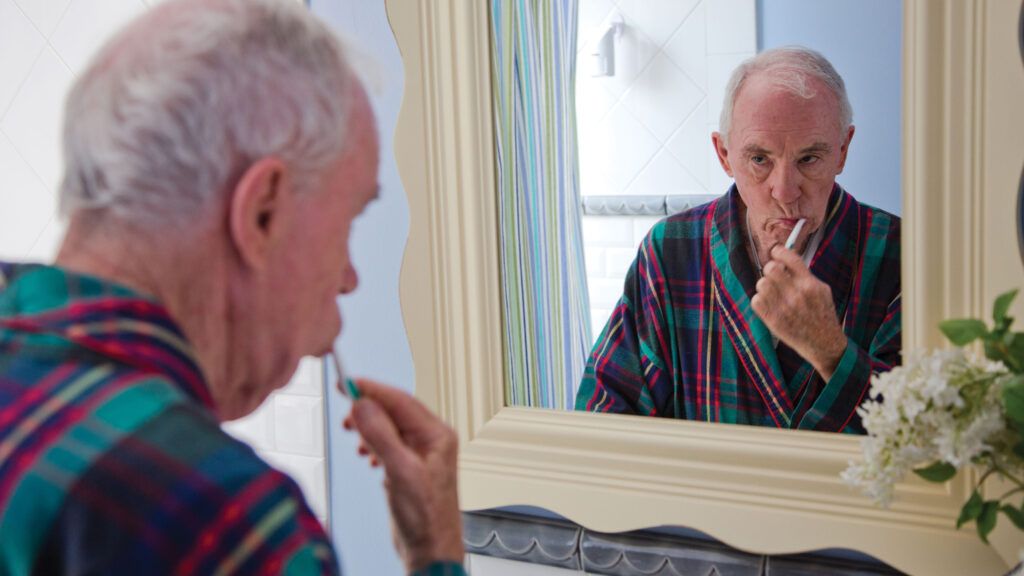 A senior citizen brushing his teeth while looking at his reflection in the bathroom mirror.