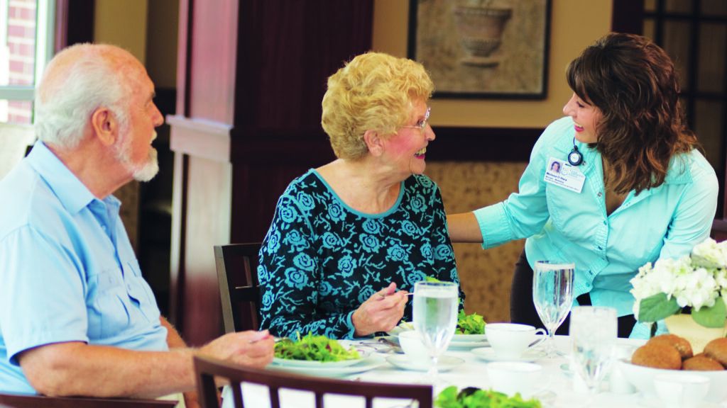 A senior citizen couple eating healthy at a table conversing with an aide.