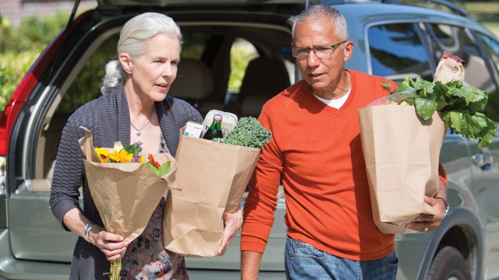 An elderly couple carrying bags of various groceries out of their car.