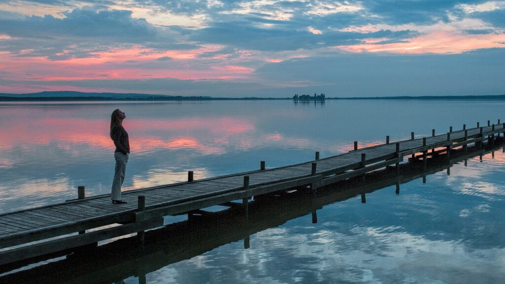 A woman gazes skyward at sunset while standing on a jetty extending into a lake