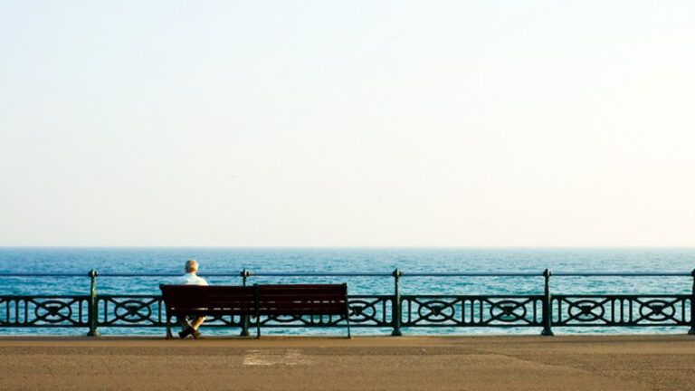 A lonely senior sitting on a bench gazing out to the sea on the beach.