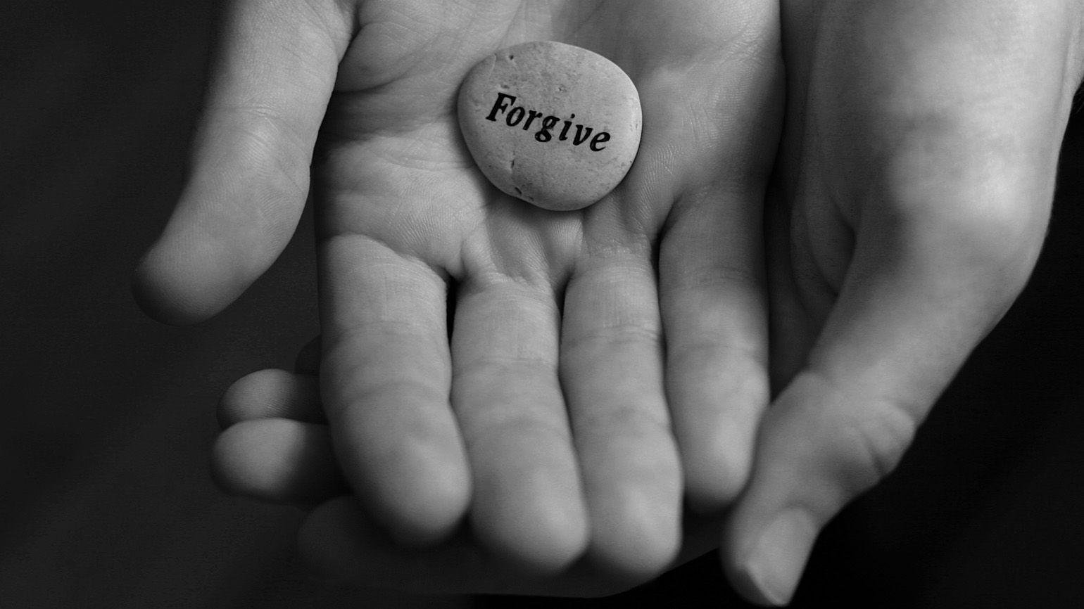 A black and white photo of hands holding a stone with 'Forgive' engraved on it.