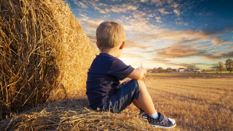 Young boy sitting on bales of straw and looking up into the sunset