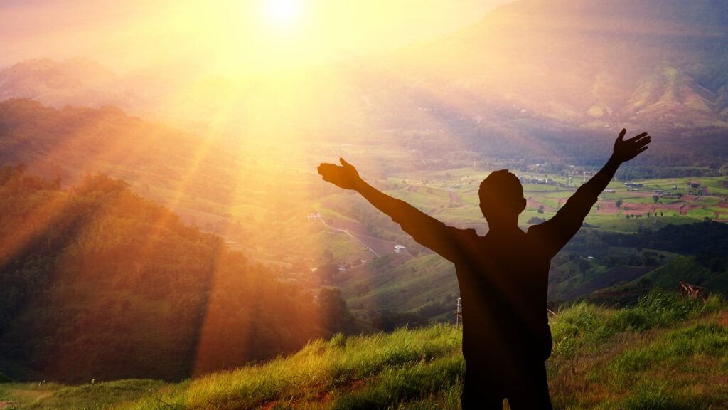 Silhouette of a man with his arms raised on a sunlit hill.