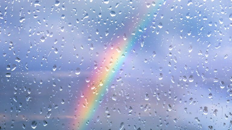 A rainbow is viewed through a window with raindrops beaded up on it