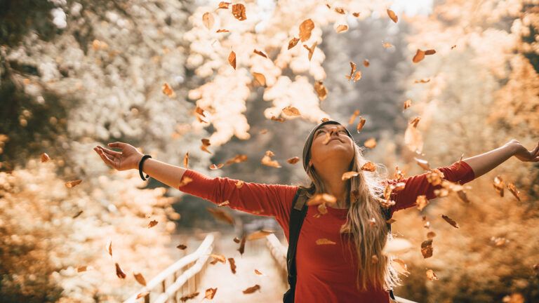 A woman stands, arms outstretched, in the falling leaves of autumn