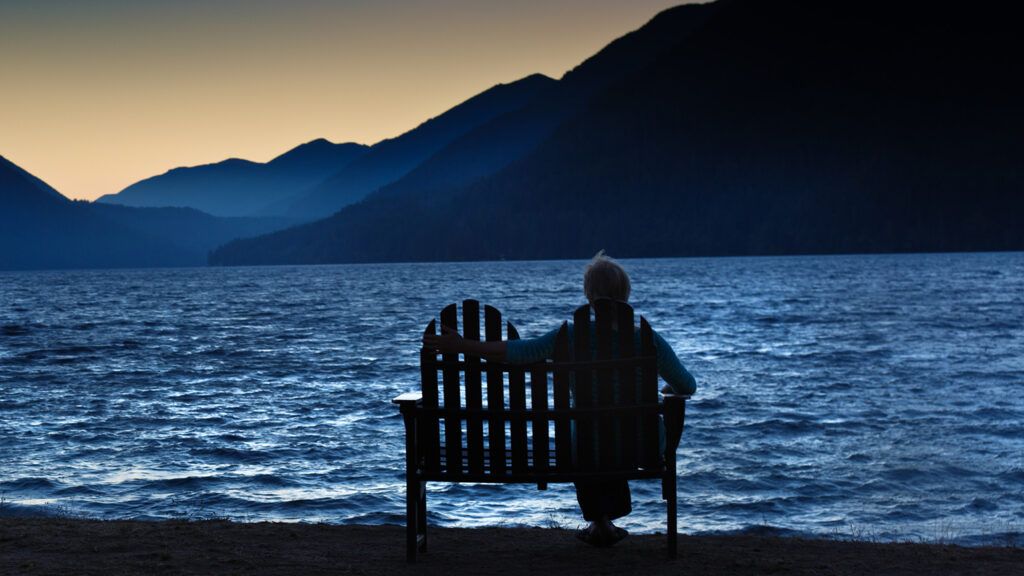 A mature woman watches the sun rise on a bench by a mountain lake