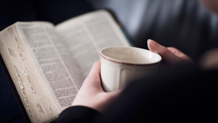 A woman's hands clasp a cup of coffee before an open Bible