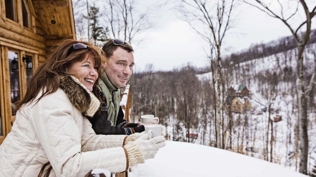 A couple looks at the snow from the balcony of their cabin with hot beverages in their hands.