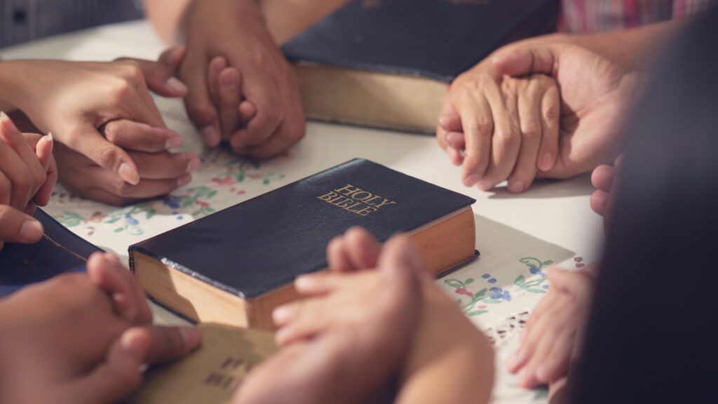 A Bible study group holding hands as a Bible rests in the center of the table.