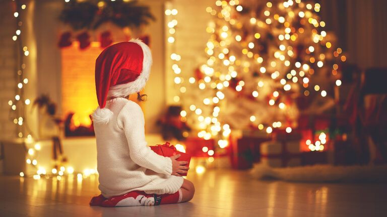A young girl in a Santa cap gazes at a Christmas tree and the presents beneath it