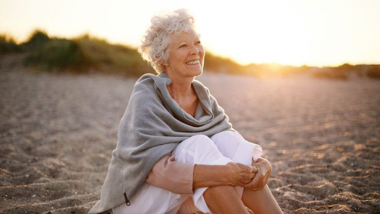 A smiling woman sits in the sand on a beach at sunrise