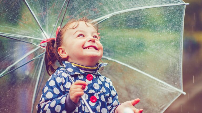 A child with an umbrella looks up at the raindrops and smiles