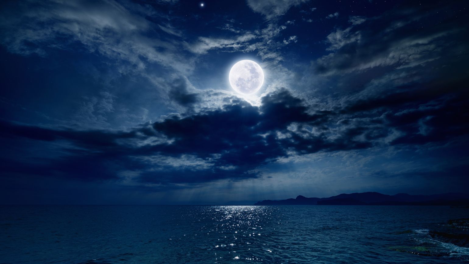 The night sky against the ocean with a full moon shining through dark clouds.